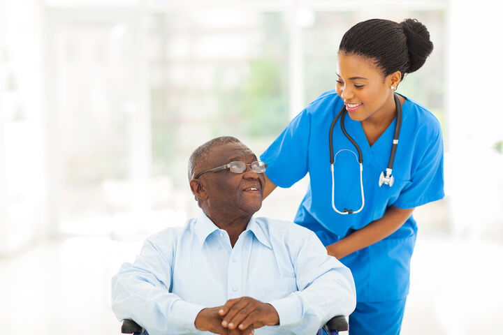 A nurse wearing blue scrubs with a stethoscope around her neck is pushing an older adult male patient in a wheelchair, while looking at him and smiling. 