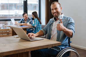 Man in wheelchair at a table, looking at a laptop and giving the thumbs up sign. 