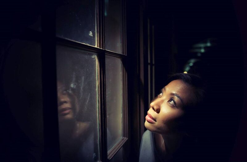 Woman w/ hopeful expression close to a window looking upwards through the window, with a bright light shining down onto her face