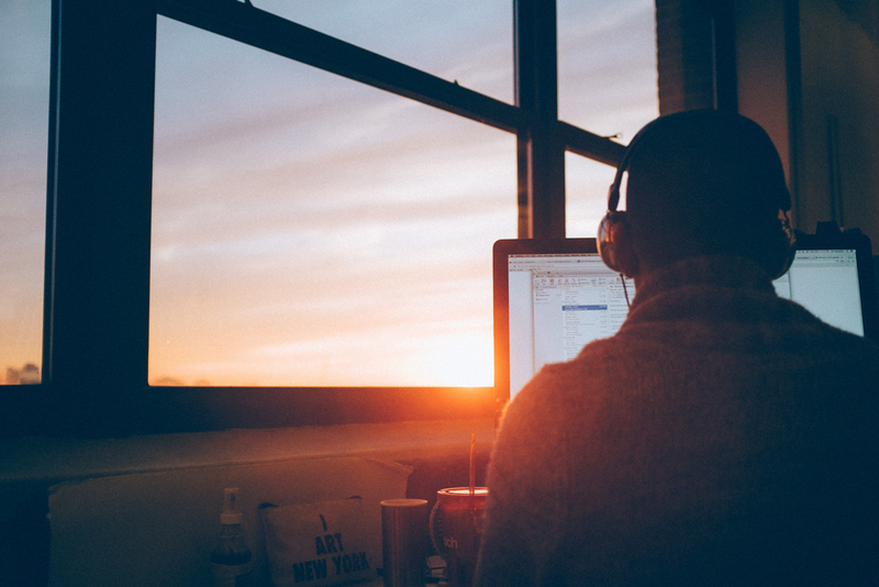 A person on a computer wearing headphones with windows displaying a sunset in the background.