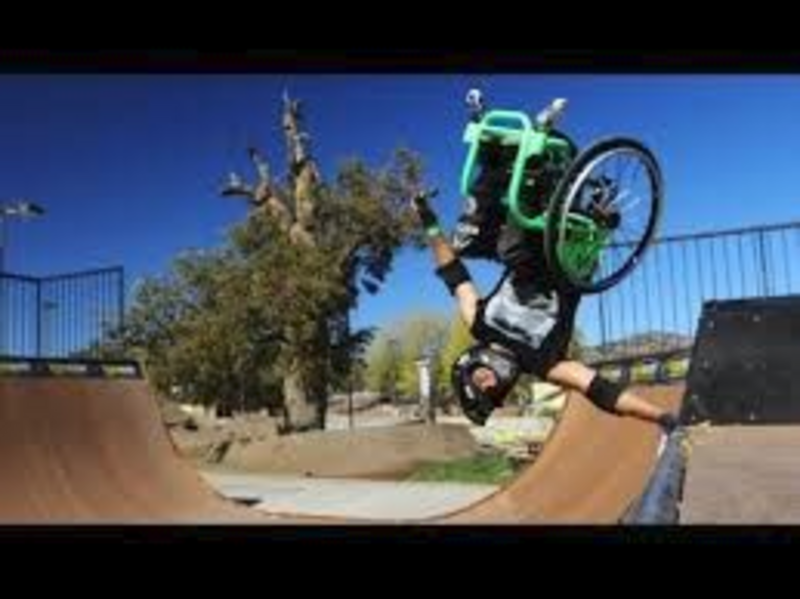 Man using a manual wheelchair on a skateboard half-pipe, mid-air and upside-down, arm extended at the top of the vertical drop-off.