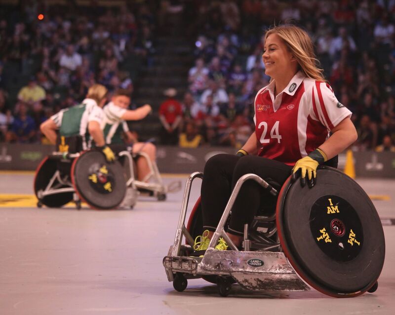 : A female wheelchair rugby player, smiling, wearing a red uniform on a court in a crowded stadium, with 2 competitors in the distance wearing green and communicating with each other.