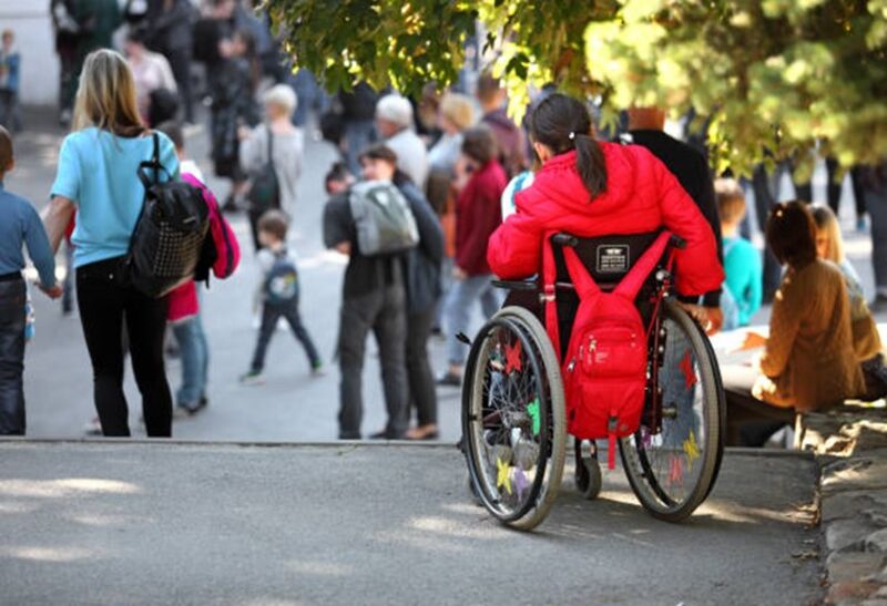a person in a wheelchair, at the top of a road, looking at a crowd of people at an outdoor venue 