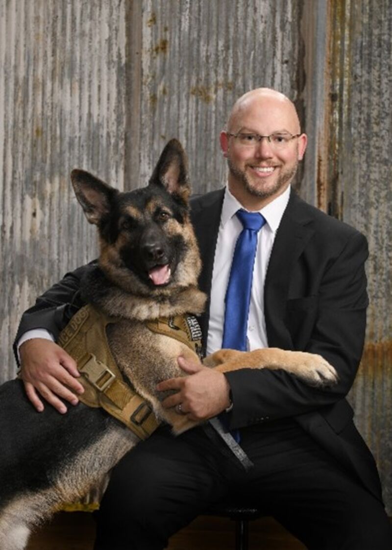 a middle aged bald white man wearing a blue suit sitting and smiling at the camera with his german shepard dog