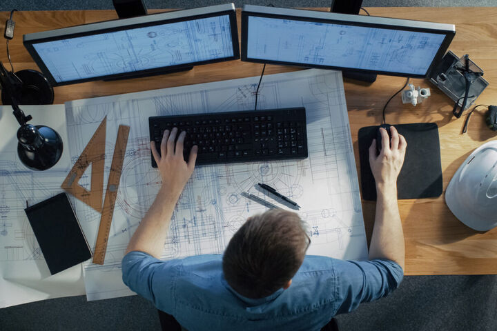 an overhead shot of an architect sitting at a draft table using computer software with two monitors and a printed schematic to work on a design