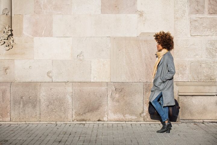 a side profile of a woman wearing a long coat walking on a sidewalk next to the face of a large building