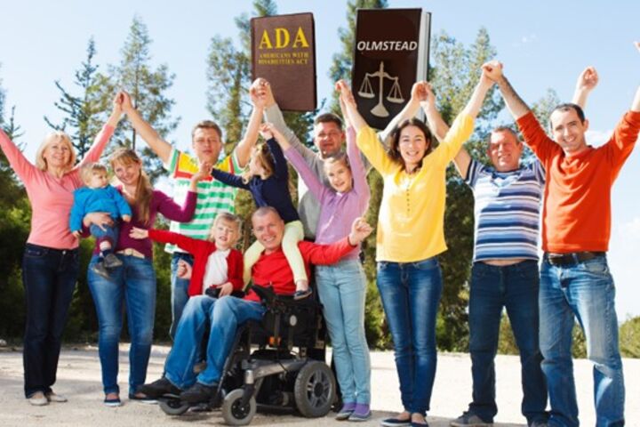 a photo of a group of 12 people, some with visible disabilities, and some without, holding their hands up with law books that say ADA and Olmstead on them, against blue sky and line of trees